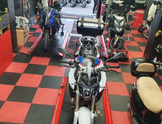 Interno officina Superbike con moto BMW bianca su ponte sollevatore, altre moto visibili, pavimento a scacchi rosso e nero. Riparazione e manutenzione moto.