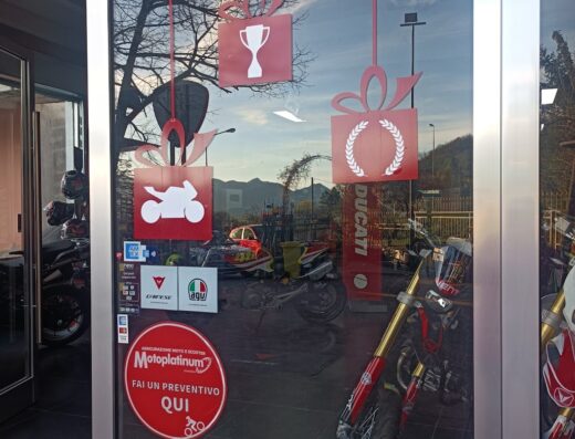 Vetrina di T.A.C. moto con adesivi decorativi a tema motociclistico, loghi Ducati, AGU, Danese, pubblicità assicurazione moto e scooter Motoplatinum, pneumatici Pirelli e riflessi di moto in vendita.
