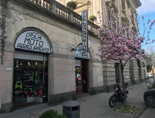 Negozio Tecnomotor s.n.c. con insegne "Caschi Moto Abbigliamento" e "Tecnomotor Ricambi Accessori", moto parcheggiata di fronte e albero fiorito.