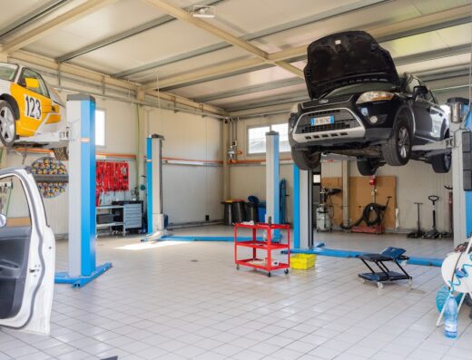 Interno di un'officina meccanica con due auto sollevate su ponti: una Ford nera con il motore in vista e un'auto da corsa gialla e bianca.