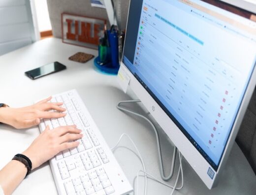 A person types on a white keyboard in front of a computer screen displaying data, with a smartphone and office supplies nearby.