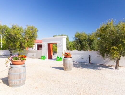 Tenuta Amostuni: ingresso caratteristico con arco bianco e porta rossa, decorato con barili di legno e vasi di fiori, bottiglie di vetro verde, immerso nel paesaggio rurale.