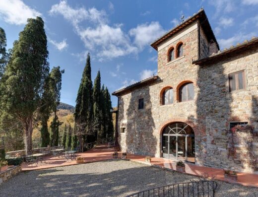 Esterno in pietra di Terre di Baccio, con cipressi, tavoli e sedie da esterno e cielo azzurro nuvoloso
