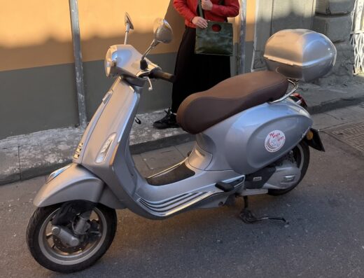Noleggio Vespa a Pisa: Vespa grigia parcheggiata in strada con bauletto posteriore e adesivo "The Vespa Rent", ideale per esplorare la Toscana.