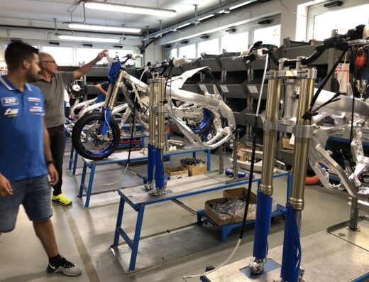 Interno dello stabilimento TM Racing SpA con operai al lavoro su telai di moto, accessori e componenti in fase di assemblaggio.