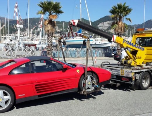 Carro attrezzi Tomaino Car Point S.R.L. che trasporta una Ferrari rossa in zona portuale con barche a vela.