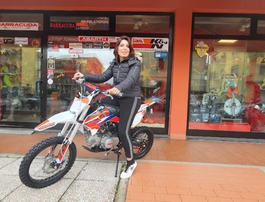 Donna seduta su una moto da cross bianca e arancione davanti al negozio Top Tuning SRL, che offre accessori e ricambi per moto.