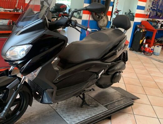 Moto scooter nera Yamaha TMAX in officina su piattaforma elevatrice per manutenzione o riparazione. Accessori moto e attrezzature visibili.