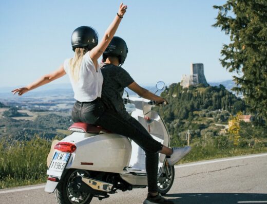 Viaggio in scooter Lambretta attraverso la Toscana: coppia con caschi esplora le colline e un antico castello. Noleggio scooter per tour unici.
