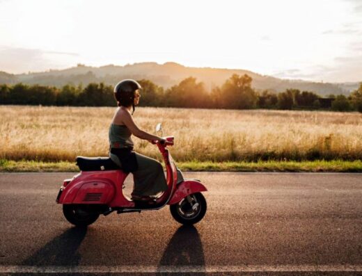 Donna che guida una Vespa rossa su una strada di campagna al tramonto. Servizio di noleggio moto e tour di Travelhoo.