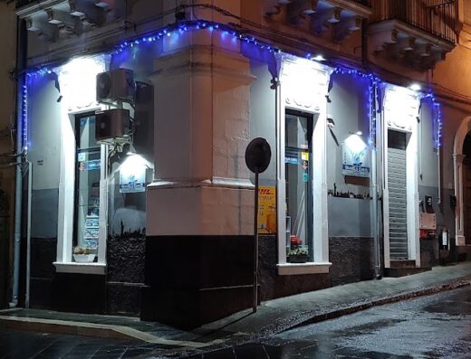 Travels Carol Agenzia Viaggi storefront at night, featuring blue Christmas lights, windows, and a street view. Travel agency services and information available here.