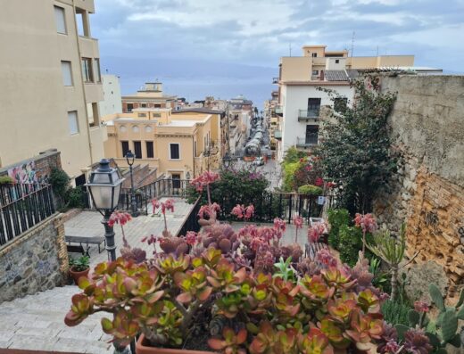 Vista panoramica di Tre Fontane, Italia, con scalinata adornata da piante grasse e fiori, che conduce verso il mare. Edifici colorati e cielo nuvoloso sullo sfondo.