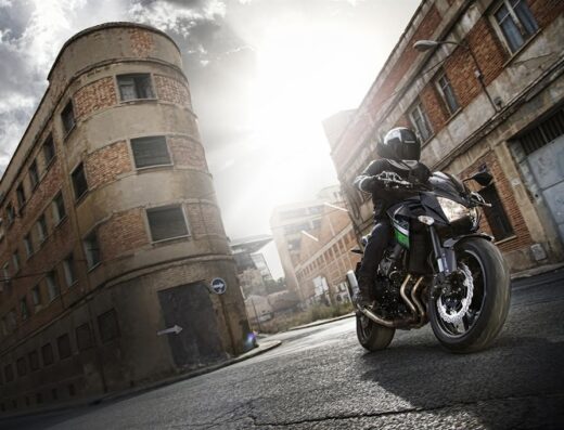 Motociclista in sella a una Kawasaki Z750 nera su strada urbana, con edifici storici sullo sfondo. Ideale per viaggi e accessori moto.