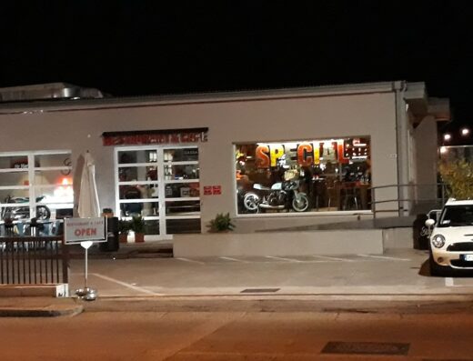 Motocicletta Triumph classica esposta all'interno di un ristorante-caffetteria a Verona, con insegna luminosa "SPECIAL" e tavoli all'aperto.