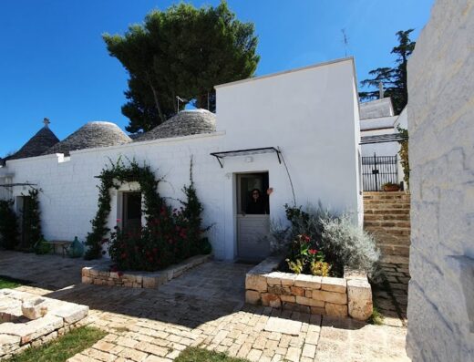 Trulli in Corte: tipica architettura pugliese con coni in pietra bianca, vegetazione curata e persona affacciata alla porta.