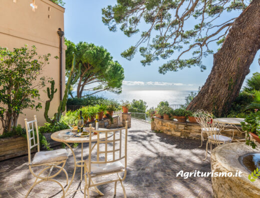 Terrazza panoramica all'Uliveto Saglietto con tavolo imbandito, sedie in ferro battuto, vista mare e vegetazione mediterranea.