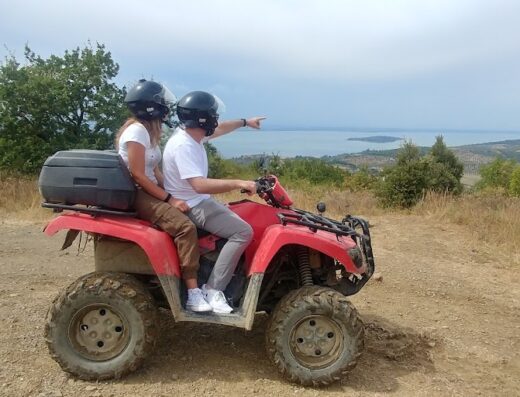 Umbria in Quad: A couple wearing helmets riding a red quad ATV on a dirt path overlooking a lake and forested hills, showcasing outdoor adventure and ATV travel in the Italian countryside.