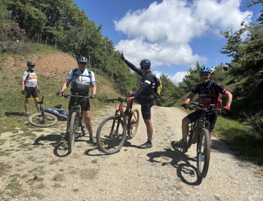 Gruppo di ciclisti in mountain bike durante una vacanza singolare, con sentiero sterrato, alberi e cielo azzurro nuvoloso.