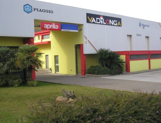 Vetrina dell'edificio Vadilonga che mostra i marchi Piaggio, Aprilia e Bianchi, un importante concessionario di motocicli e rivenditore di accessori