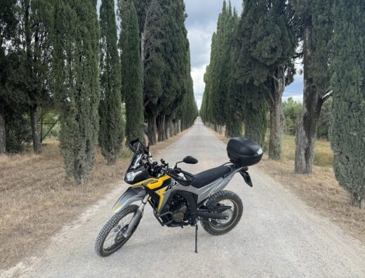 Una moto da enduro grigia e gialla di Pastrimotors parcheggiata su una strada sterrata in Val d'Orcia, fiancheggiata da cipressi.