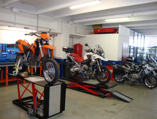 Officina Vallauto: diverse moto, tra cui una KTM arancione su un ponte sollevatore, e BMW GS bianche e nere, pronte per assistenza e riparazione.