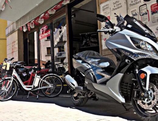 Vallillo Moto: Scooter Kymco argentato in esposizione davanti al negozio di Vallillo Nicola, con biciclette parcheggiate e insegna Kymco visibile.
