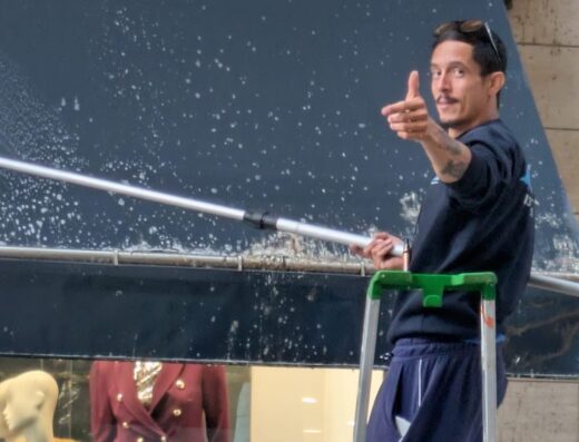 Un uomo su una scala esegue la pulizia e sanificazione a vapore di una vetrina con abbigliamento esposto, utilizzando un'asta con spazzola.