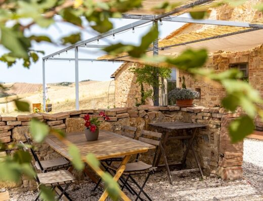 Patio esterno di una casa in pietra nella Val d'Orcia, con tavolo in legno, sedie pieghevoli e vista sulle colline toscane