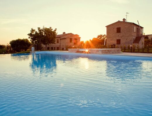 Piscina al tramonto presso Verde Benessere Villaggio Agrituristico, con riflessi del sole sull'acqua e casali sullo sfondo.