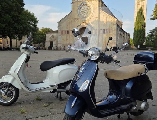 Due scooter Vespa parcheggiati in una piazza di Verona, con una chiesa storica sullo sfondo. Ideale per esplorare la città su due ruote.