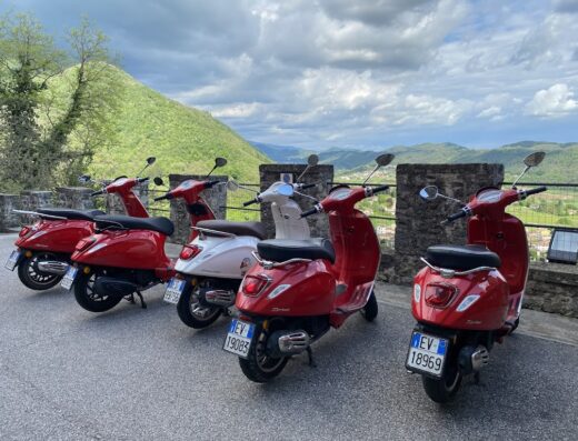 Noleggio Vespa Dolomiti: Diverse Vespa rosse e una bianca parcheggiate con vista panoramica sulle montagne, ideali per un tour avventura.