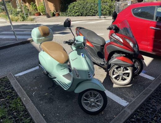 Noleggio Vespa e scooter a Salò by Garda Detailing: una Vespa verde acqua con bauletto e sella marrone parcheggiata vicino a uno scooter a tre ruote rosso.