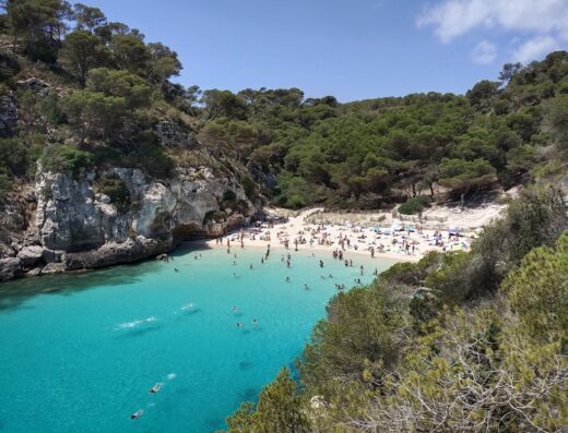 Vista panoramica di Cala Macarella a Minorca, con spiaggia affollata, acqua turchese cristallina, vegetazione mediterranea e cielo azzurro. Ideale per viaggi e vacanze.