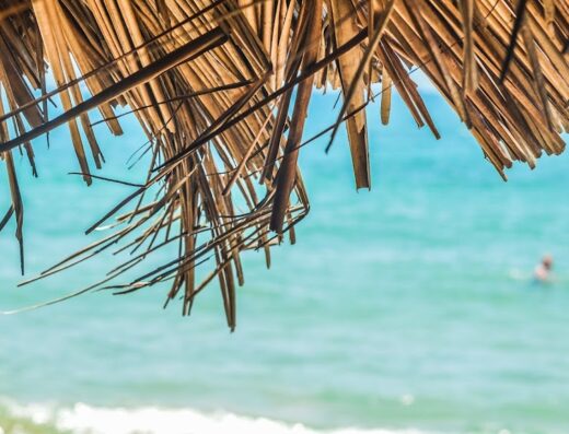 Dettaglio di un ombrellone di paglia con vista sul mare turchese, persona che nuota sullo sfondo. Viaggi Manuzzi.