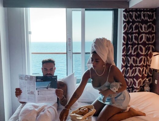 Una coppia si gode la colazione a letto in una cabina di crociera con vista sull'oceano; l'uomo legge il giornale e la donna tiene in braccio un peluche.