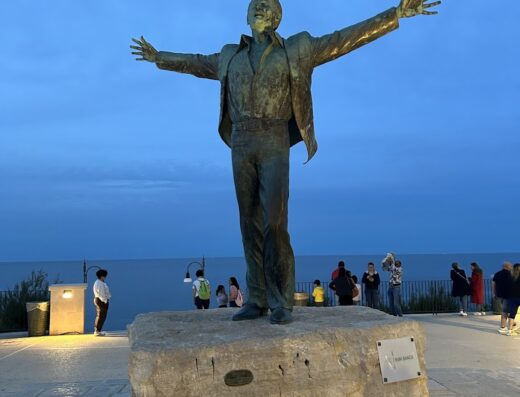 Statua in bronzo di Domenico Modugno a Polignano a Mare, Puglia, con il mare sullo sfondo. Località turistica ideale per viaggi in moto e accessori.