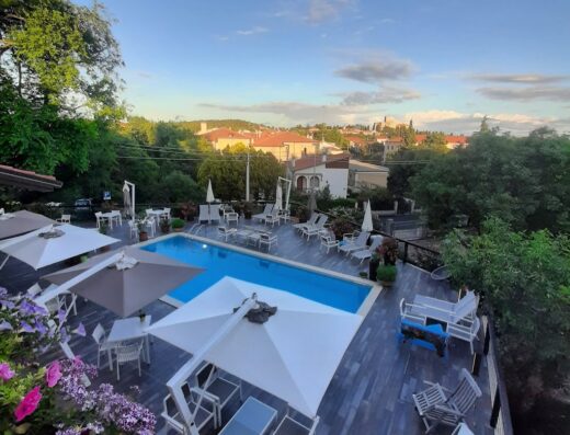 Veduta aerea della Villa Borgo Duino con piscina, lettini prendisole, ombrelloni, tavoli e sedie. Sullo sfondo, case con tetti rossi e vegetazione lussureggiante.