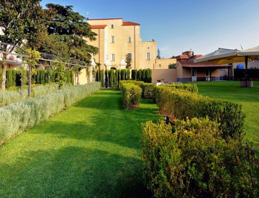 Green lawn at Villa Buonanno with trimmed hedges, trees, and the villa building in the background on a sunny day.