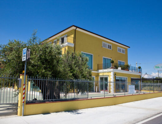 Villa Imago, una struttura ricettiva gialla con balconi e recinzione, immersa nel verde e ideale per viaggiatori in moto che cercano un soggiorno confortevole e servizi dedicati al turismo su due ruote.