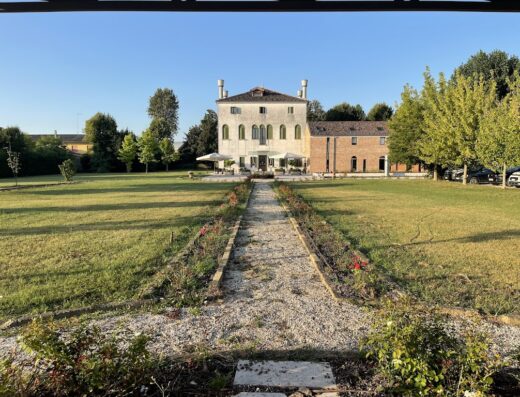 Villa Martin Hotel exterior view with garden, gravel path lined with roses, lawn, outdoor seating, and attached red brick building.