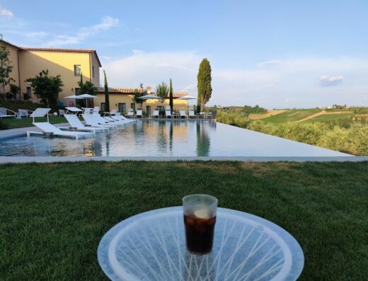 Villa Petriolo infinity pool view with a drink on a small table, sun loungers and Tuscan landscape in the background.