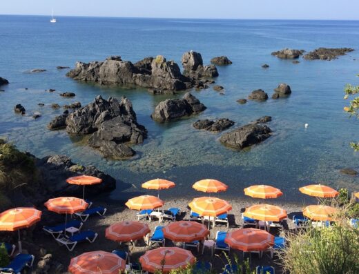 Vista panoramica sulla spiaggia di Illicini con ombrelloni arancioni, lettini blu, scogli e mare cristallino, ideale per vacanze e relax.