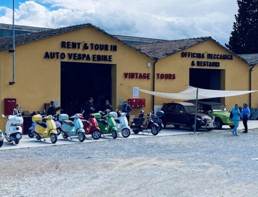 Noleggio Vespa e tour in Toscana: fila di Vespa colorate e auto d'epoca davanti all'officina Vintage Tours, per un'esperienza di viaggio unica su due ruote.
