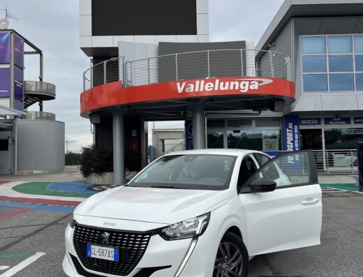 Peugeot 208 bianca parcheggiata di fronte all'Autodromo di Vallelunga, con una portiera aperta, ideale per il noleggio auto a Cassino.