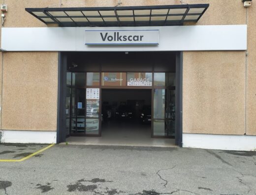 Ingresso di Volkscar Sas, concessionaria e officina autorizzata Volkswagen. Insegna con logo Volkswagen e scritta Volkscar ben visibile sopra l'entrata del garage con auto all'interno.