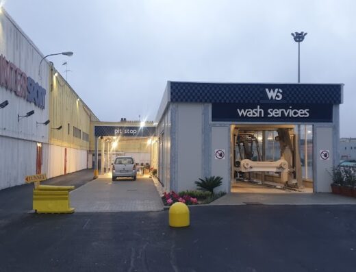 Servizio di lavaggio moto Wash Services S.R.L.: auto bianca in fila per il lavaggio automatico, insegna "Pit Stop" e struttura esterna ben illuminata.