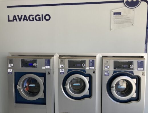 Lavanderia self-service con tre lavatrici Electrolux grigie e blu disposte in fila, con indicazioni numeriche e cartello informativo.