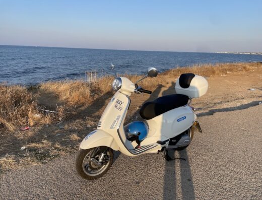Una Vespa bianca parcheggiata vicino alla costa con l'oceano sullo sfondo. Accessori per moto come bauletto posteriore e casco visibili.