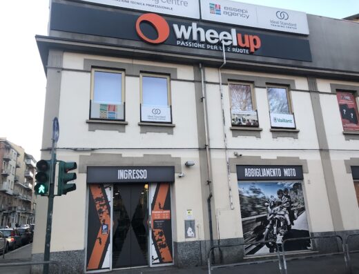 Immagine della facciata del negozio Wheelup, specializzato in accessori e abbigliamento moto. Si vede l'ingresso e un cartellone pubblicitario di abbigliamento moto.