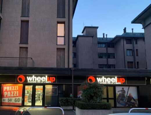 Negozio Wheelup con vetrine illuminate che mostrano offerte su accessori e abbigliamento per moto. Insegna luminosa con logo Wheelup e la scritta 'Passione per le 2 ruote'. Orari di apertura visibili.
